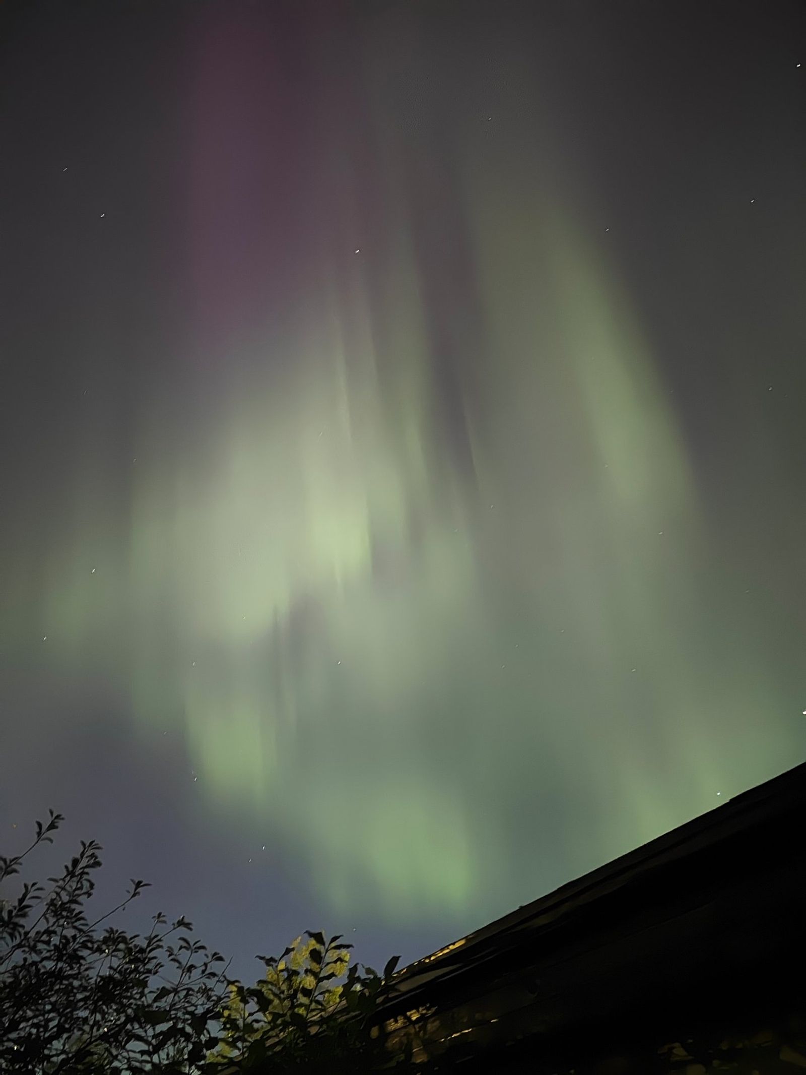 Pystysuora kuva värikkäistä revontulista, alla osa rakennuksen kattoa.