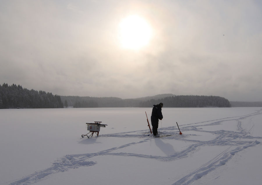 Kalastaja lumen peittämän järven päällä auringon paistaessa taivaalla pilvien takana